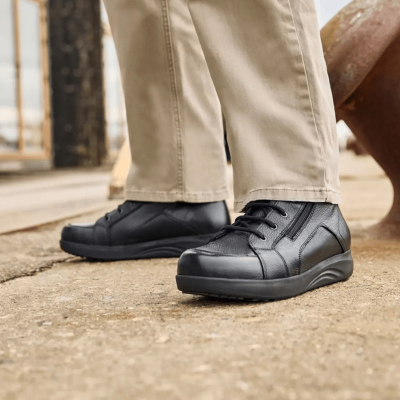 Un homme en pantalon beige et bottines New Feet noires pour homme debout sur une surface grise