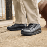 Un homme en pantalon beige et bottines New Feet noires pour homme debout sur une surface grise