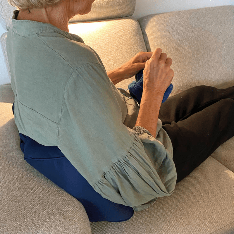 Senior assis sur un canapé avec un coussin lombaire bleu pour le soutien du dos, tricotant et se relaxant